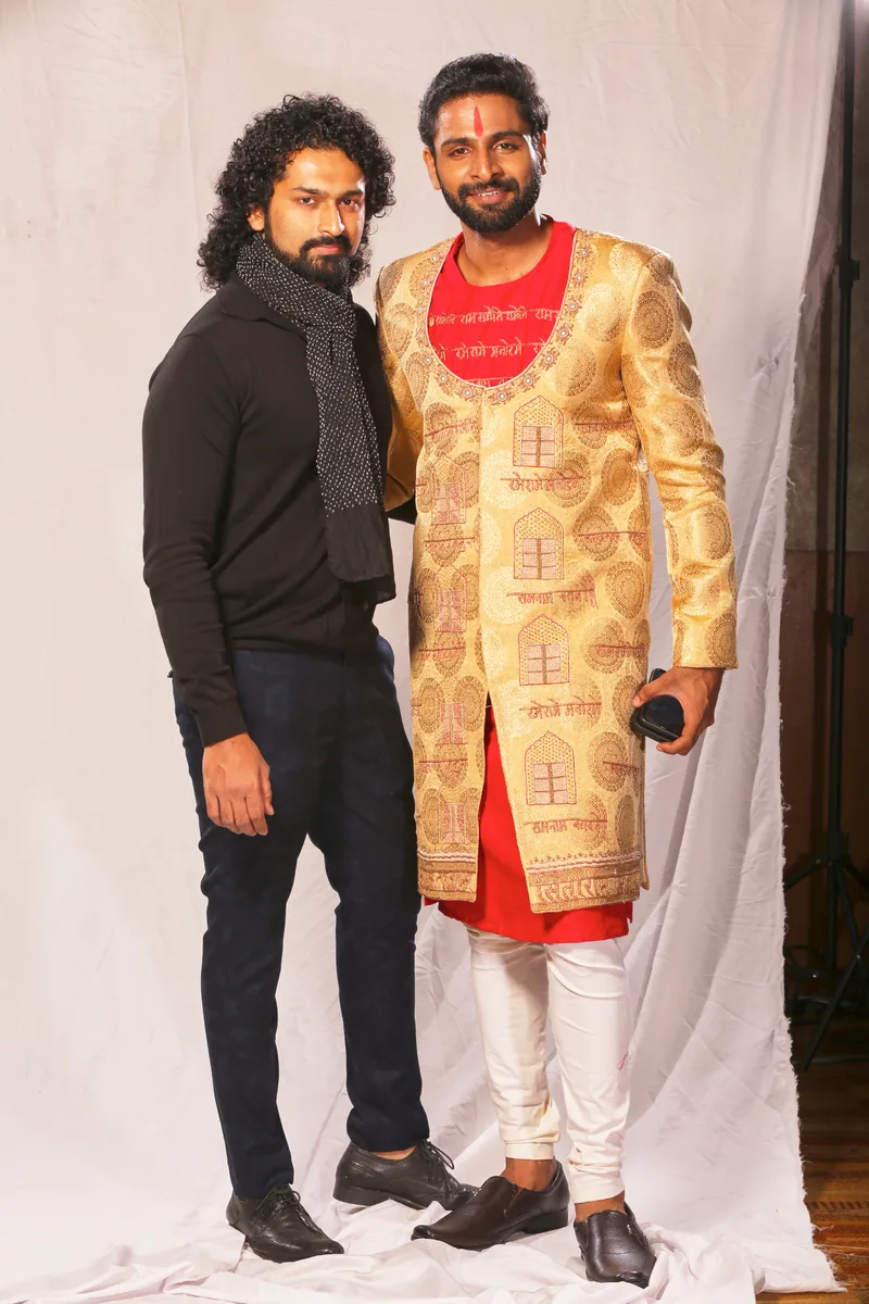 Two men posing backstage beside a gold sherwani with red details