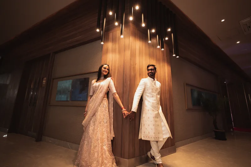 Couple holding hands in ivory and blush wedding outfits beneath pendant lights