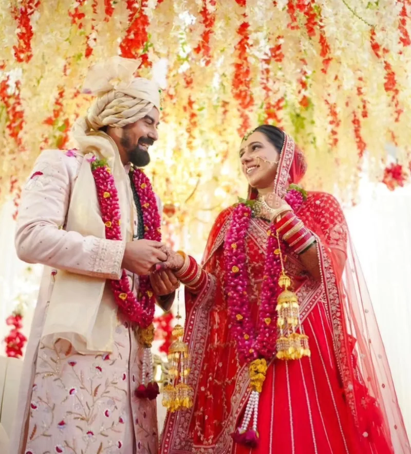 Bride in a red sari and groom in an ivory floral sherwani exchanging smiles