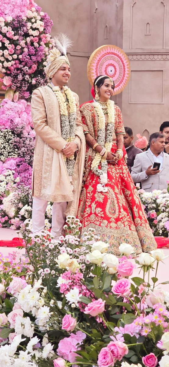Bride and groom in red and ivory wedding outfits surrounded by pink flowers